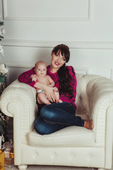 Happy young mother with bare newborn baby sitting in armchair in decorated room for Christmas