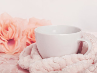 Big white mug with coffee or tea tucked with a soft blanket next to roses on pink marble desk. Copy space