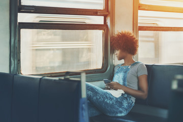 Fototapeta na wymiar Thoughtful young cute African American female is sitting alone inside of moving suburban train and looking outside the window on the train coming from the opposite direction and holding smartphone
