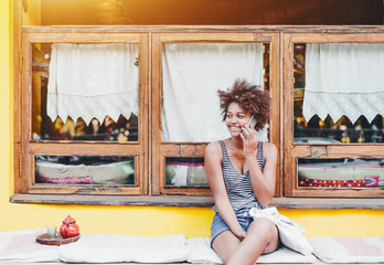 Laughing cute Brazilian girl speaking on smartphone while sitting on cushion in front of yellow wall and window; young smiling African American female with cellphone waiting friend in street cafe
