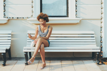 Charming young black girl is sending online message via smartphone while sitting on street bench with facade building behind her with copy space zone for text, your advert or other information - Powered by Adobe
