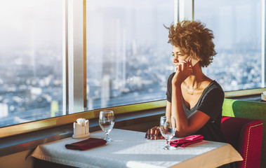 Charming pensive young black female is sitting at the table of modern luxury restaurant located on...