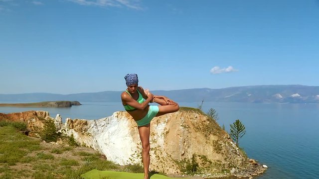 Young brunette professional yoga woman practices yoga moves outdoors near the giant blue lake baikal.