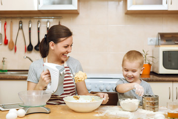 Obraz na płótnie Canvas Little kid boy helps mother to cook Christmas ginger biscuit in light kitchen with tablet on the table. Happy family mom 30-35 years and child 2-3 in weekend morning at home. Relationship concept