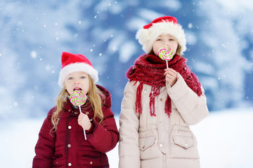Two adorable little sisters wearing Santa hats having huge striped Christmas lollipops on beautiful winter day