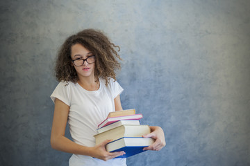 Curly hair teen girl with glasses stands next to the wall and holds several books