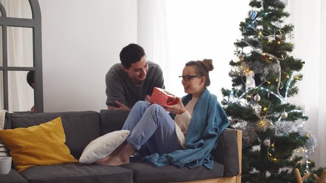 Man giving surprise christmas gift for woman sitting on sofa.