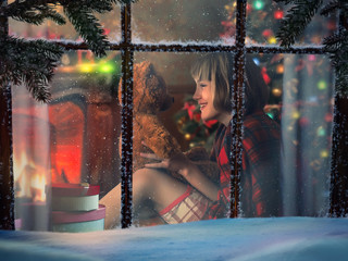 Looking through snowy window at home. Happy child with Teddy bear. Inside the house warm, fireplace. Happy Christmas
