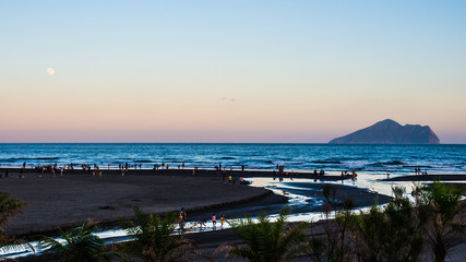 Obraz premium The tourists are enjoyed with black sand and pacific sea of Waiao beach, Taiwan. The sun is setting with vanilla sky, the moon is also showed up on the left side above horizon and the isolated island