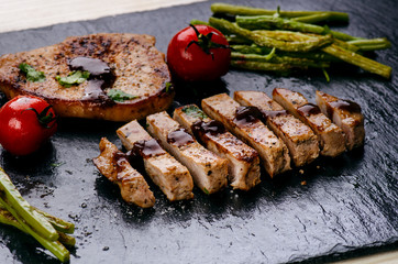 Grilled pork chops, steaks with vegetables, tomatoes, beans and sauce on a black slate. Fresh meat with foam. Dark background. Grilled and barbecue concept. 