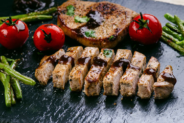 Grilled pork chops, steaks with vegetables, tomatoes, beans and sauce on a black slate. Fresh meat with foam. Dark background. Grilled and barbecue concept. 
