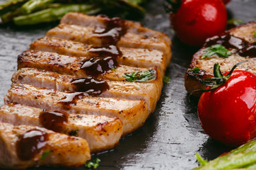 Grilled pork chops, steaks with vegetables, tomatoes, beans and sauce on a black slate. Fresh meat with foam. Dark background. Grilled and barbecue concept. 
