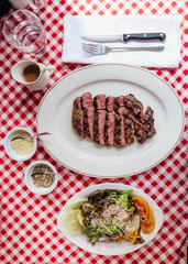 Top view of Sliced medium rare charcoal grilled wagyu Ribeye steak in white plate on red and white pattern tablecloth with seasoning, BBQ sauce and cutlery.