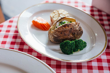 Baked potato served with grilled carrot, corn and broccoli in white plate on red and white pattern tablecloth.