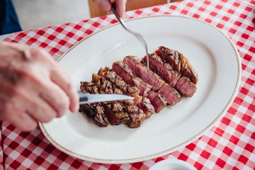Sliced medium rare charcoal grilled wagyu Ribeye steak in white plate on red and white pattern tablecloth.
