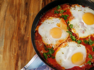 A pan of fried eggs with tomato sauce and parsley on a wooden background. Shakshuka a traditional meal of the Jewish cuisine