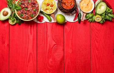 Vegetarian Mexican food concept: refried black and red beans. guacamole, salsa, chili, tortilla chips and fresh ingredients over vintage red rustic wooden background. Top view, flat lay