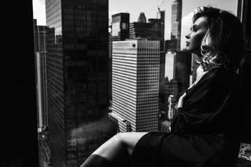 Seductive young woman in black silk robe lies with naked shoulders before a panoramic window in New York