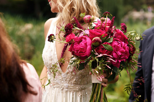 Pretty bride holds rich dark pink bouquet of peonies in her arm