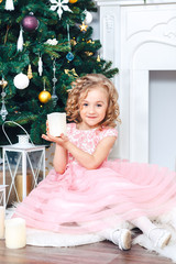 a small child sits under a Christmas tree in a beautiful pink dress, holds a candle, around gifts