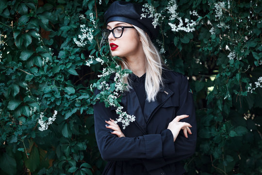 Fashion close up portrait of stylish young blond woman with red lips, trendy glasses and leather black hat in grape leaves. Autumn trendy look.  High fashion concept.