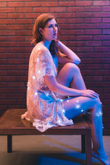 Young caucasian woman in sheer dress poses with string lights against a brick wall