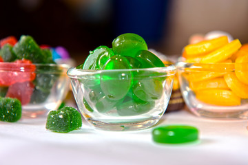 A glass bowl filled with glossy green hard candies, surrounded by other colorful sweets including yellow candies and sugar-coated gummies, arranged on a white surface for a vibrant candy display