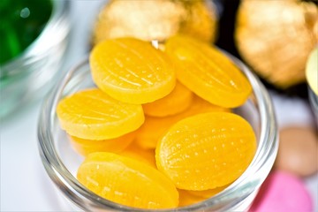 A glass bowl filled with vibrant yellow hard candies with a textured surface. The bright candies are glossy and oval-shaped, placed in a clear bowl with golden-wrapped chocolates in the background