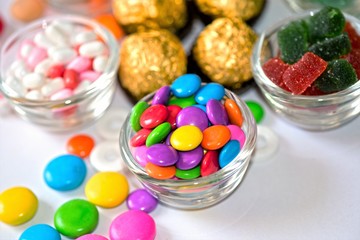 A vibrant assortment of colorful candy-coated chocolate gems in a glass bowl. These glossy, round candies come in a mix of bright colors, including red, blue, green, yellow, orange, and purple, making