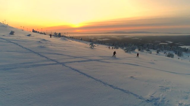 AERIAL Group of unrecognizable skiers skiing down the snowy mountain slope at winter sunset. People skiing fresh powder snow at golden sunrise in Levi, Finland. Winter activity on sunny winter morning