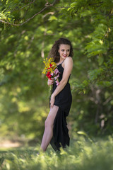 Pretty young lady wear black long dress with deep neckline stands under a tree holding bouquet of wild flowers