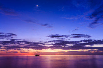 Silhouette of fishing boat at sunset with the beam of sun shines on the surface of the sea and moon in the sky