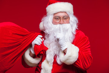 Portrait of Santa Claus with huge red sack keeping forefinger by his mouth and looking at camera