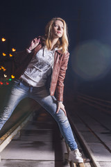 Youth Lifestyle Concept and Ideas. Happy Smiling Caucasian Woman In Leather Jacket and Blue Jeans Playing on Rails With Bag Outdoors at Night.Halogen and Flash Lights are Used.