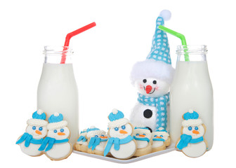 Snowmen sugar cookies with blue scarfs and fluffy blue and white hats on a plate with bottles of milk green and red straws. More snowmen cookies standing next to glasses of milk