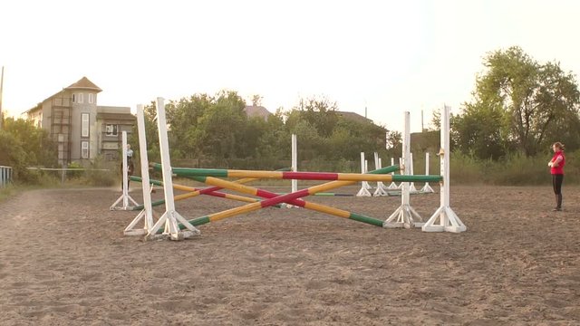 Professional rider on a horse jumps over a barrier. Competitive rider training jumping over obstacles at sunset. Mare jumps over two barriers in training at the racetrack. Slow motion.