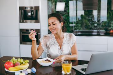 Portrait of beautiful happy woman working with laptop while breakfast with cereals and milk. Eating at home.