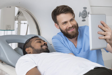 Radiologist Showing Digital Tablet To Patient Lying On CT Scanne