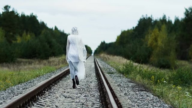 The view from the back. The young woman with make-up of dead bride for Halloween dressed in white wedding gown going away along the railway among the autumn forest. 4K