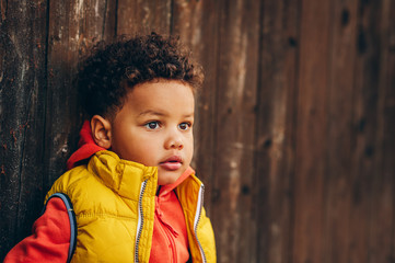 Outdoor portrait of adorable toddler boy posing outside against brown wooden background, wearing orange hoody jacket any bright yellow vest coat