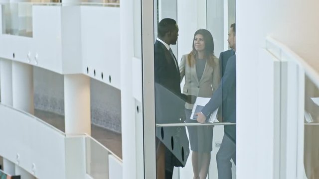Multi-ethnic business team riding down and discussing something in modern glass elevator in office building