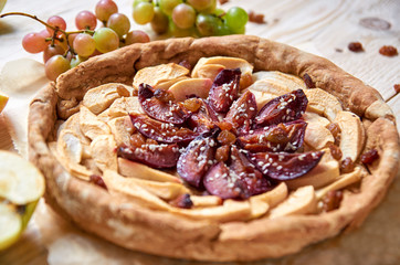 A homemade apple plums pie decorated with fresh apples, grapes, brown raisins and sesame on light wooden background. Close up view