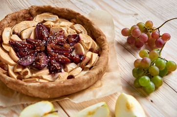 A homemade apple plums pie decorated with fresh grapes, apples and sesame on light wooden background. Side view