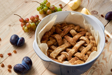 Just baked homemade apple pie in blue baking form decorated with fresh apples, grapes and brown raisins on light wooden background. General view