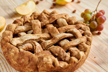 A homemade apple pie decorated with fresh sliced apples, grapes and brown raisins on light wooden background. Side view