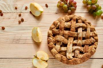 A homemade apple pie decorated with fresh apples, grapes and raisins on light wooden background. Top view