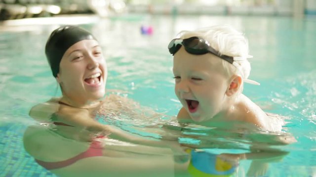 Happy little boy is swimming in the pool together with his mother. She is holding him and teaching how to swim. Happy family spending time together