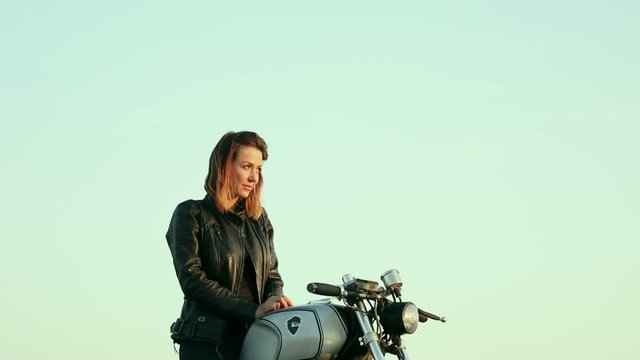 Sexy biker girl wearing leather jacket sitting on custom build motorcycle cafe racer and looking at the sunset. Toned