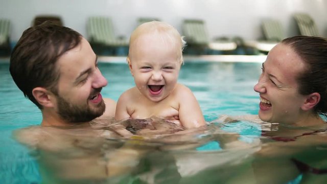 Young mother and father are holding their cute child in the swimming pool and kissing each other in their lips. Happy child is smiling
