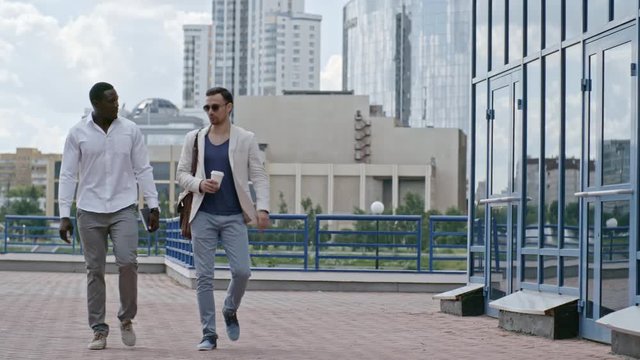 Tracking of business partners walking together towards the camera at summer day and chatting; African man holding organizer and Caucasian businessman going with coffee cup